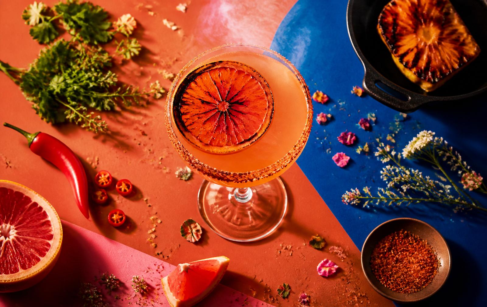 Editorial food-magazine flat lay: a smoky Shawnarita cocktail with charred grapefruit and chili salt rim, herbs, peppers, and citrus on bold terracotta and cobalt paper.